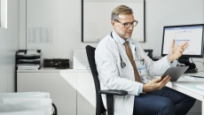 Healthcare provider sitting in a chair at a desk with a computer on it while looking at a tablet