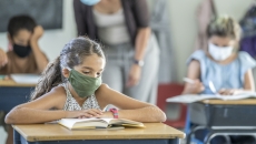 12 year old girl wearing a reusable, protective face mask in classroom while working on school work at her desk.