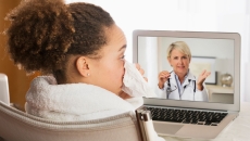 Person sitting in a chair while blowing their nose and talking to a healthcare provider on a computer