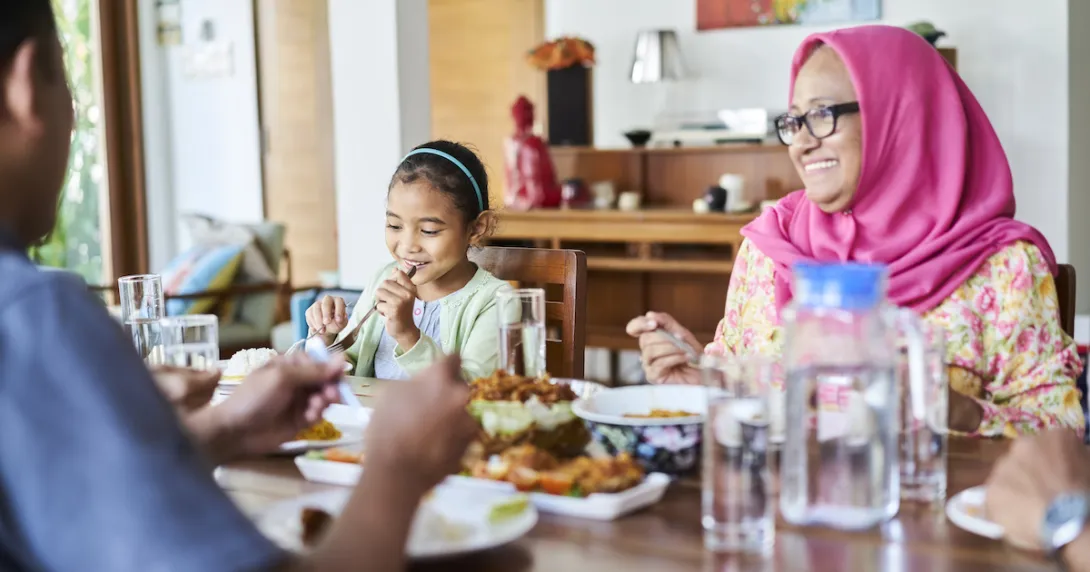 Three people sitting around a table with food on it. One person is wearing a pink head scarf, one a blue shirt and the other a white shirt