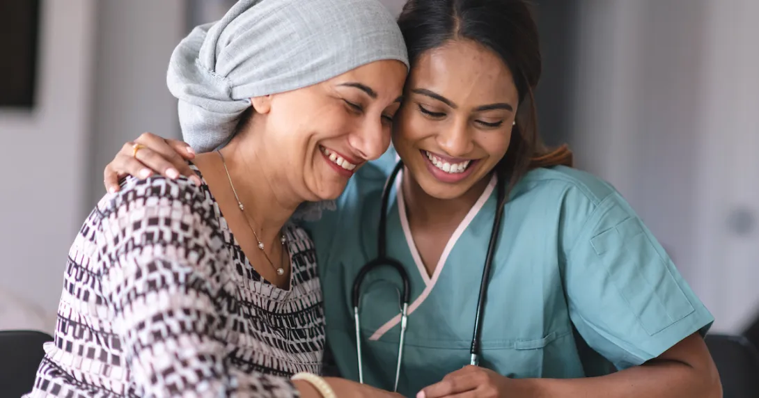 A healthcare provider with scrubs on sitting next to an oncology patient wearing a head scarf