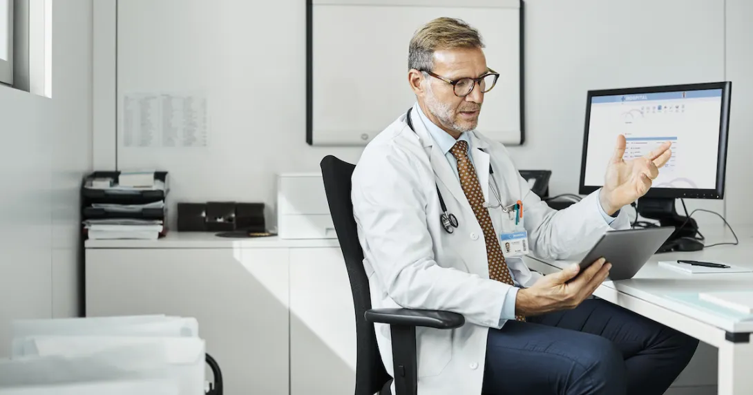 Healthcare provider sitting in a chair by a desk with a computer on it while talking to someone on a tablet
