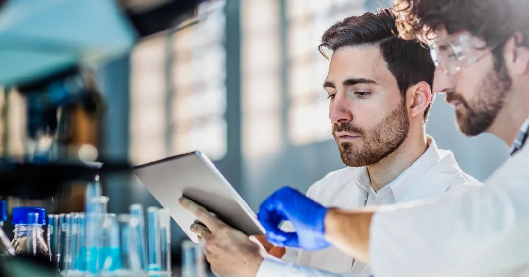 Two scientists in a laboratory setting looking at a tablet while surrounded by beakers