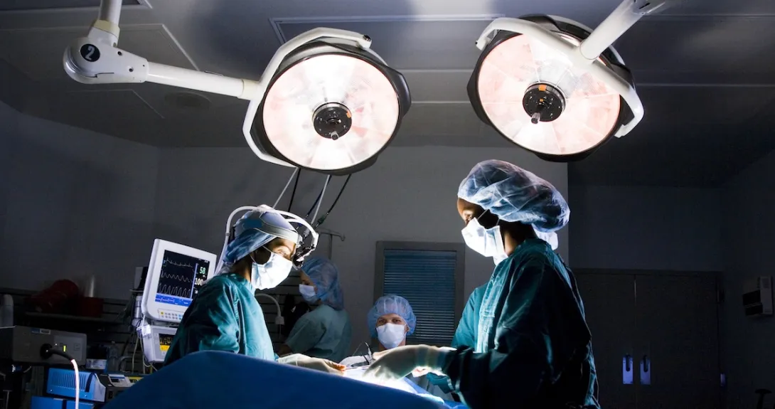 Healthcare providers in an operating room looking down at a patient on a table with large lights above them