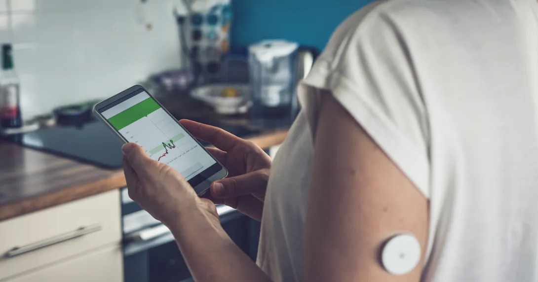 A close up of a person wearing a CGM while tracking glucose levels on their phone