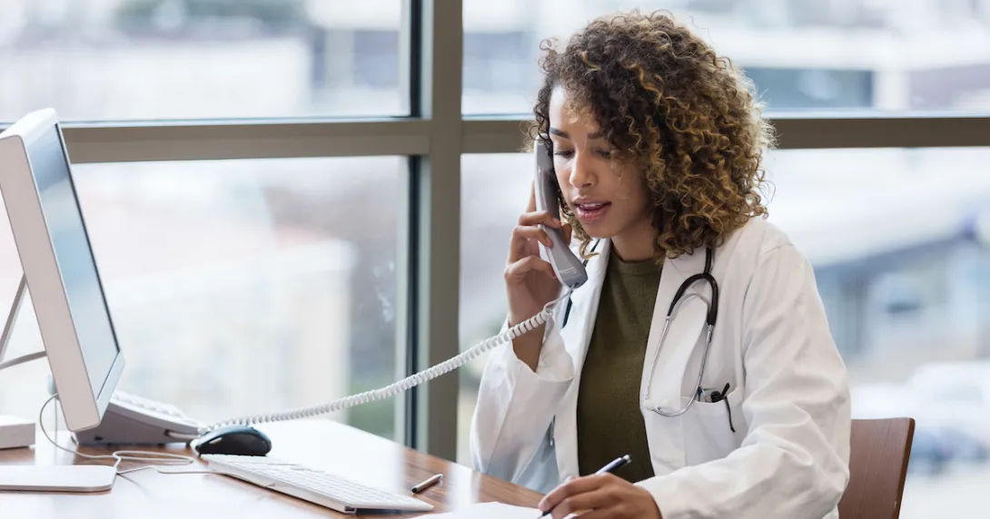 A doctor, sitting in her office, uses the landline to make phone calls regarding patient records.