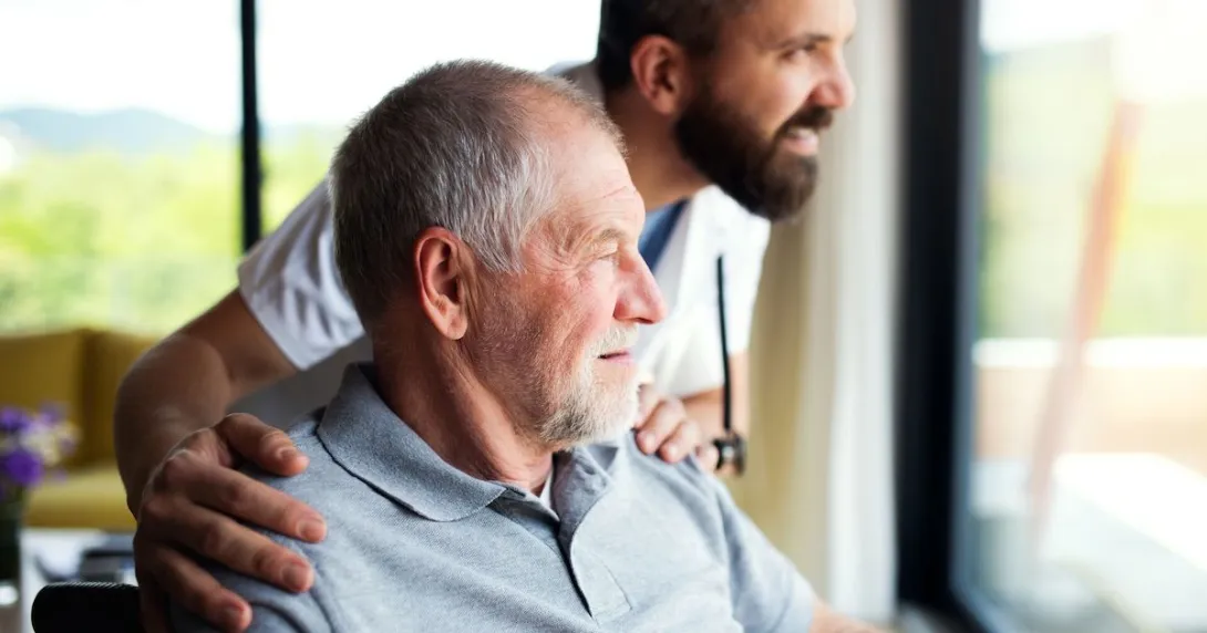 A person in a wheelchair and a healthcare provider standing beside them looking out a window