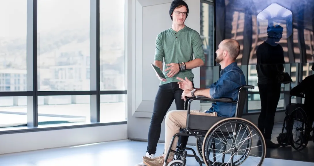 Person in a wheelchair and another people standing beside them in a room with a large window