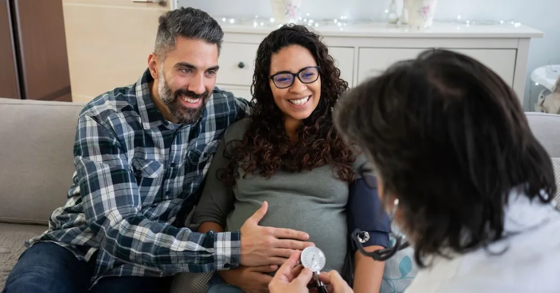 Two people sitting on a couch, one pregnant and the other touching their stomach, with another person sitting across from them