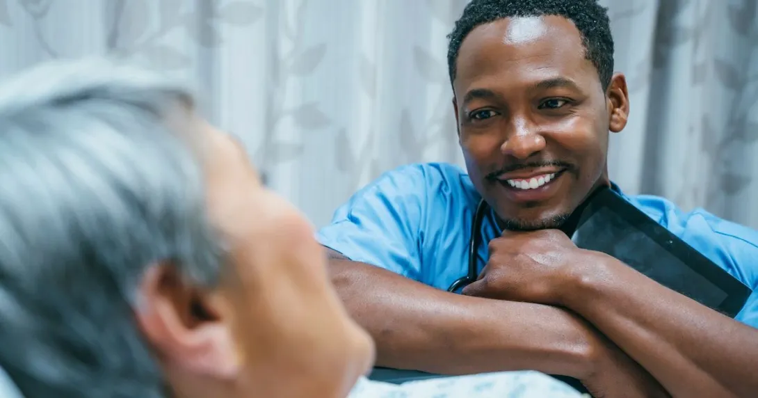Healthcare provider sitting beside a patient's bed with the patient in it while the provider is holding a tablet