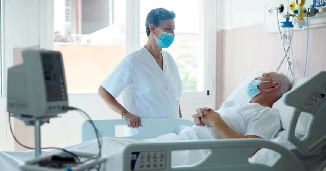 Person in mask standing over person in hospital bed